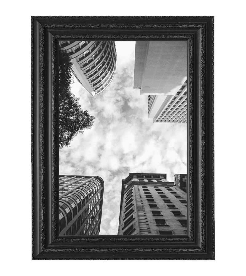 Black-and-white photo of tall buildings shot from below, framed by a black ornate frame—perfect for showcasing in any space. Discover elegant photo frames Mississauga residents love, with a cloudy sky visible between the structures.
