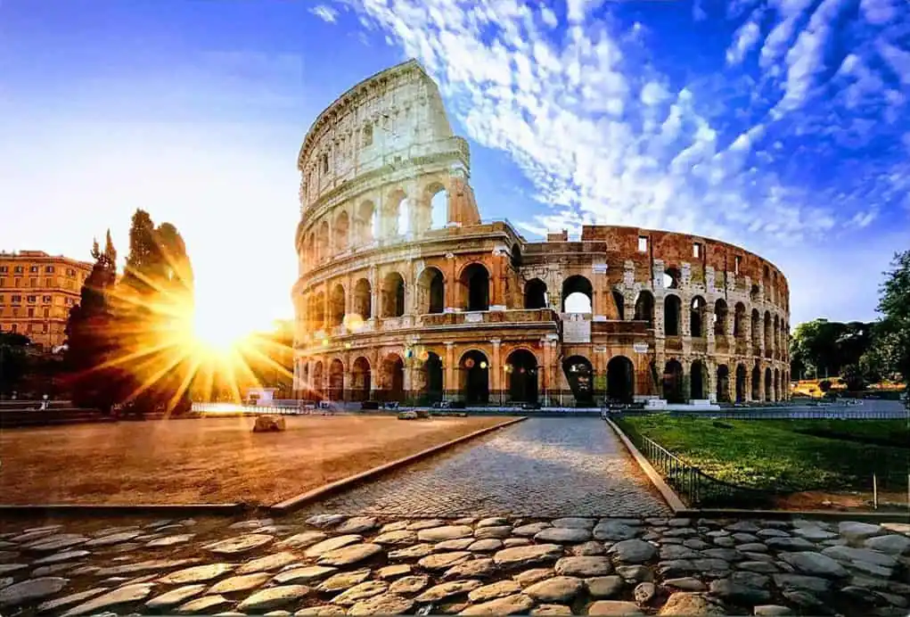 The Colosseum in Rome at sunrise, captured by supreme pictures, showcases a bright sunburst on the left and a clear blue sky above.